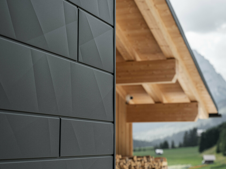 Close-up van de Siding.X gevelpanelen in P.10 zwart van PREFA aan de Casa Hoffe in Sappada, Italië. Het gevelsysteem van aluminium is vakkundig aangebracht in een modern, abstract ontwerp dat een aantrekkelijk contrast vormt met de zichtbare houten elementen van het dakgebinte. De innovatieve geveloplossing past naadloos in de alpine omgeving en staat voor de combinatie van functionaliteit, duurzaamheid en esthetische bouwcultuur.