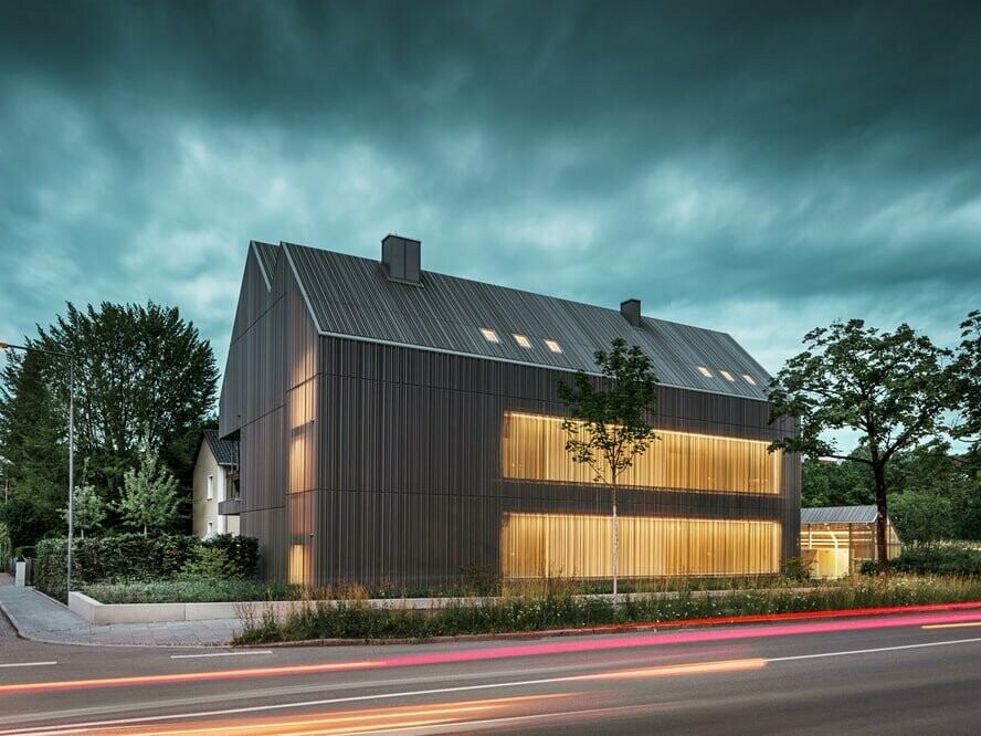 Das Mehrfamilienhaus in Dachau in der Dämmerung. Durch die teilweise gelochte Fassade kommt das Licht vom Stiegenhaus durch und verleiht dem Ganzen einen außergewöhnlichen Look.