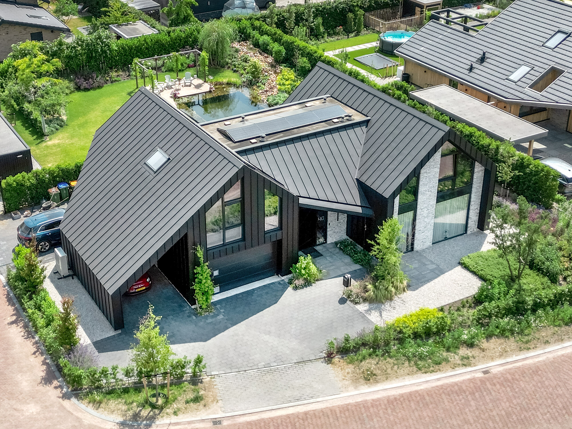 Luchtfoto van een moderne eengezinswoning met een steil, zwart Prefalz zadeldak met ingewerkte zonnepanelen. De architectuur combineert grote glaspartijen met geometrische vormen. Een carport is in het gebouw geïntegreerd. De villa beschikt over een goed onderhouden tuin met vijver en terras, en is gelegen in een rustige buurt in voorstedelijk gebied.