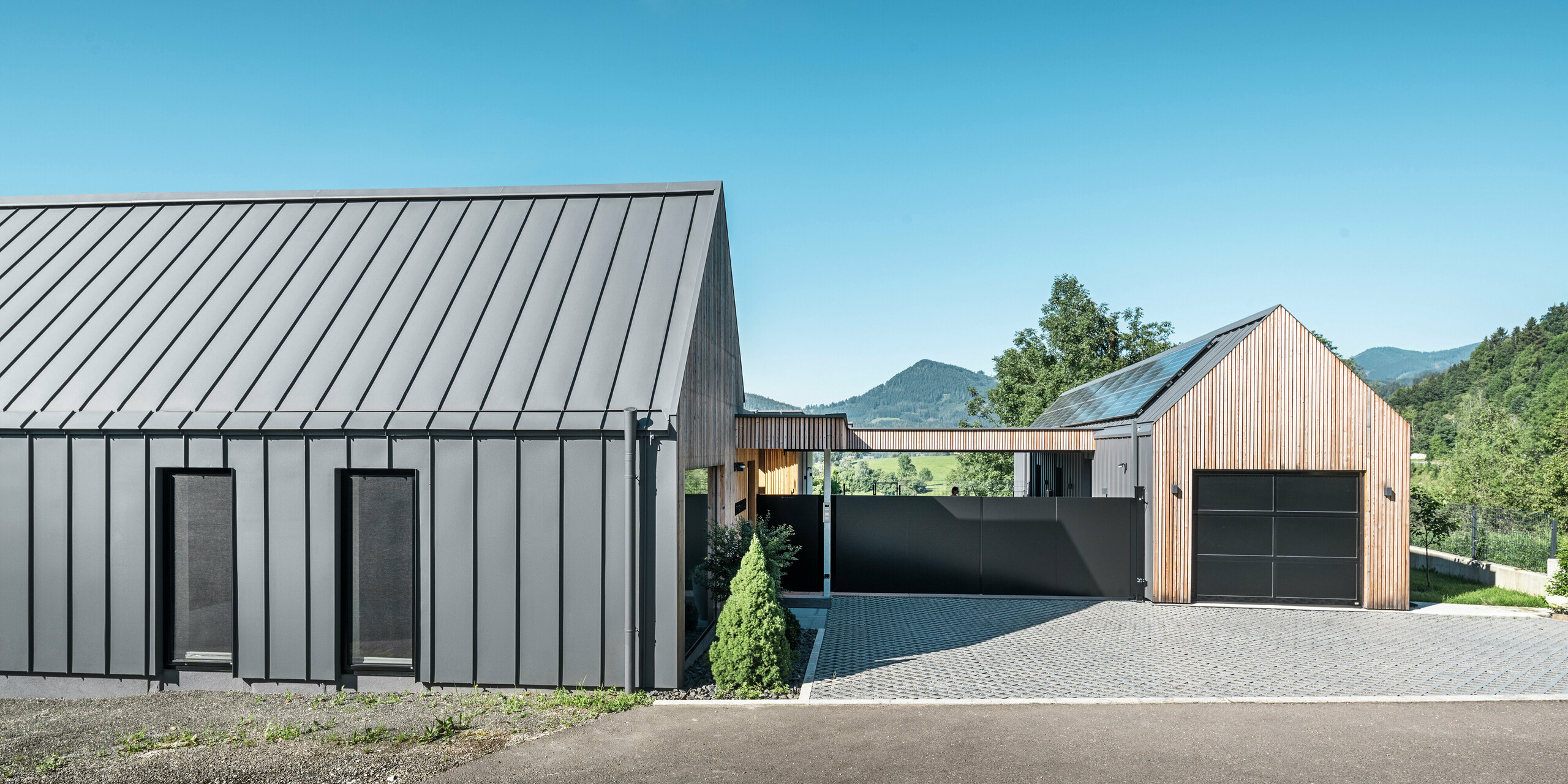 Eingangsbereich eines Einfamilienhauses mit angrenzender Garage in Pogier, Österreich, inmitten einer malerischen Landschaft. Dach und Fassade beider Gebäude sind mit langlebigem PREFALZ in P.10 dunkelgrau verkleidet, was ein modernes Erscheinungsbild erzeugt. Die Garage verfügt über eine harmonisch integrierte Holzverkleidung, die sich nahtlos in die umliegende Landschaft einfügt. Der gepflasterte Hof und die klaren Linien des Entwurfs ergänzen die Ästhetik des Grundstücks, während im Hintergrund die sanften Hügel das Bild einer friedlichen grünen Oase im grünen Herzen Österreichs vervollständigen.