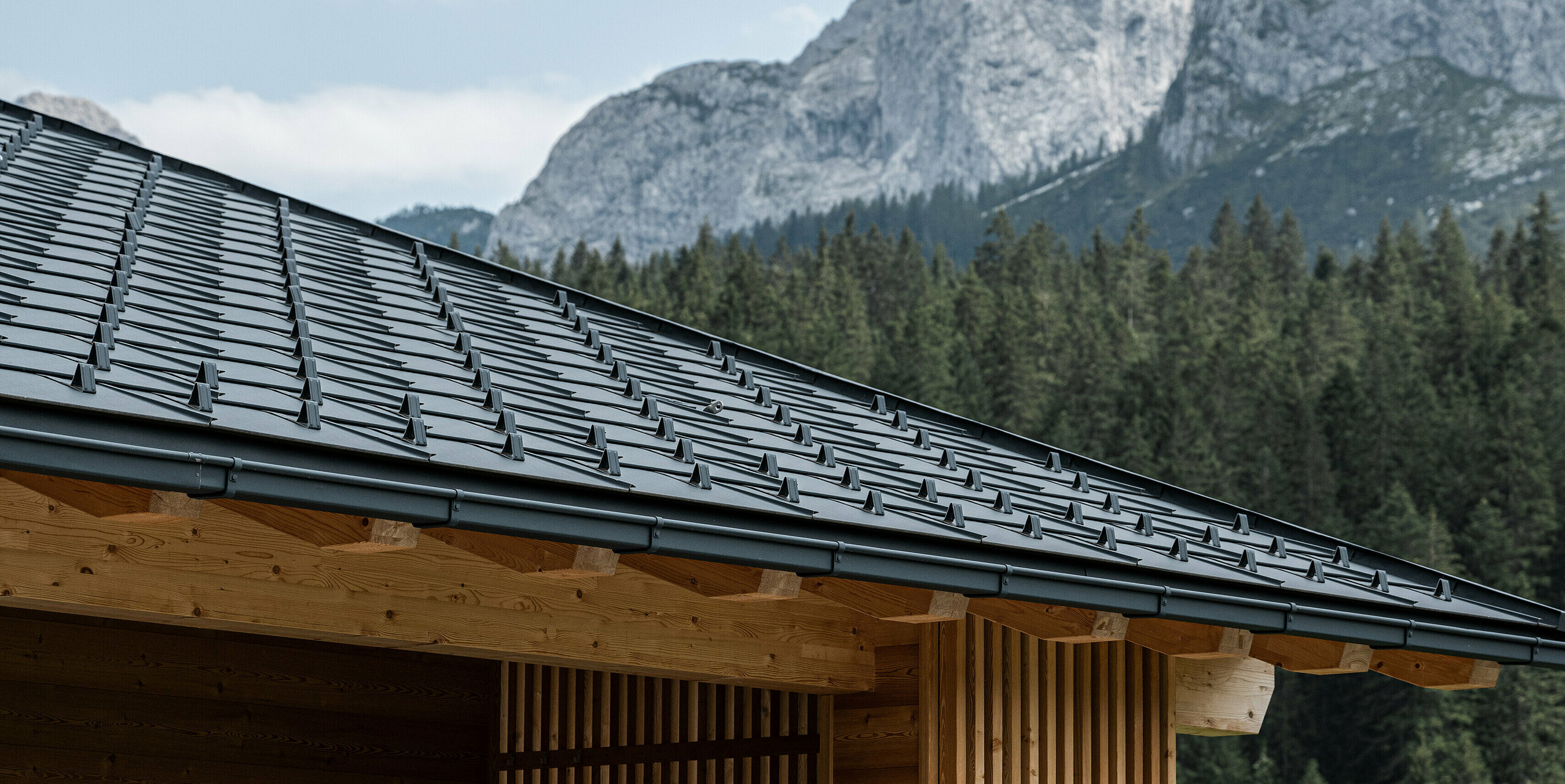 Seitenansicht der Loggia von Casa Hoffe in Sappada, Italien, mit einem Dach aus PREFA Dachplatten R.16 in P.10 Schwarz. Die sorgfältig arrangierten Dachplatten ergänzen die natürliche Holzstruktur der Loggia und bieten einen modernen Touch im Herzen der Dolomiten. Das blühende Blumenarrangement auf dem Balkon und die beeindruckenden Berge im Hintergrund runden das Bild eines idyllischen Alpenzuhauses ab.