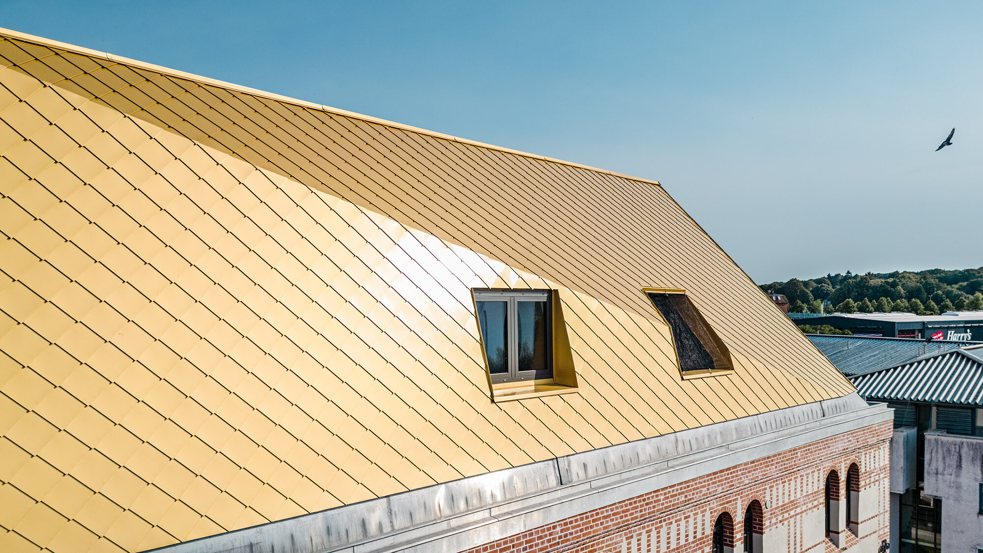 Das Dach mit den mayagoldenen Aluminiumrauten und einem Knick aus der Nähe, ein Teil der denkmalgeschützten Fassade ist sichtbar. 