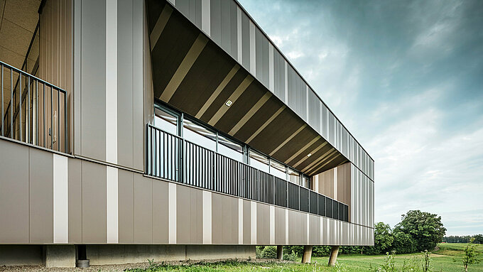 Detailansicht der modernen Aluminiumfassade des Veranstaltungszentrums ZIB Nikolai in St. Veit, mit vertikalen Streifen in Braun- und Beigegrau-Tönen. Die Loggia mit schwarzen Geländern und großzügigen Fenstern bietet einen Blick ins Grüne, während die Fassade durch ihre klaren Linien und geometrischen Formen besticht. Im Hintergrund ist eine natürliche Landschaft unter einem leicht bewölkten Himmel zu sehen.