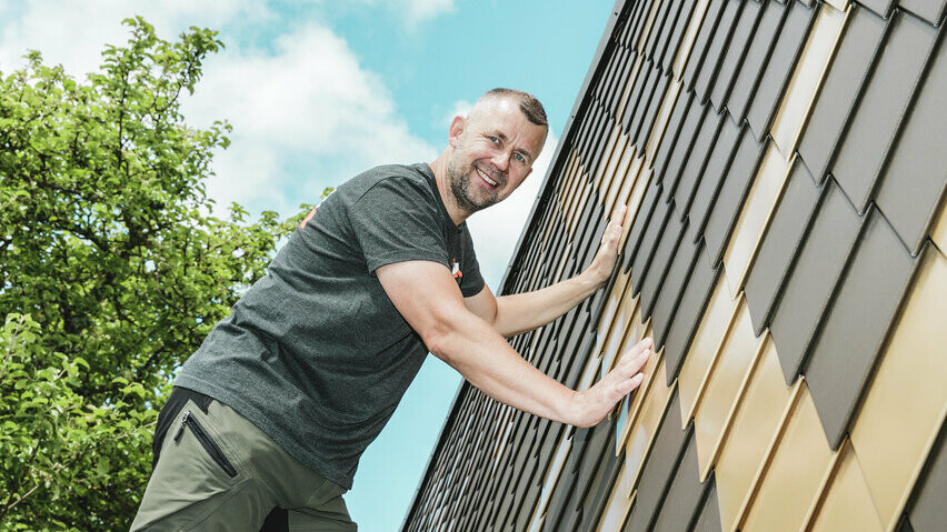 PREFA Handwerker mit grauem T-Shirt an einer PREFA Rauten-Fassade in Braun und Mayagold.