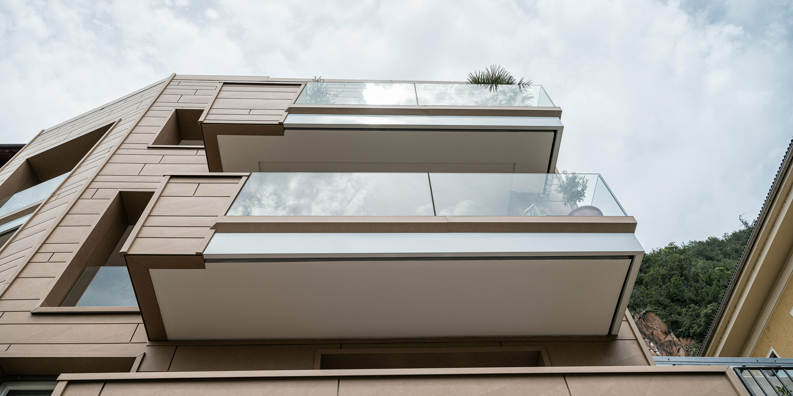 Blick von unten auf die Villa Laura in Italien, verkleidet mit PREFA Siding.X in Sandbraun, welche eine robuste Eleganz ausstrahlt. Die klaren, modernen Linien des Gebäudes kontrastieren mit dem weichen Himmel und den natürlichen Hintergründen, wobei die Fassade durch ihre Struktur und Farbe eine eindrucksvolle Verbindung zum natürlichen Ambiente schafft. Das durchdachte Design vermittelt einen Eindruck von Offenheit und Leichtigkeit und spiegelt den innovativen Einsatz von Materialien im Einklang mit der Umgebung wider.