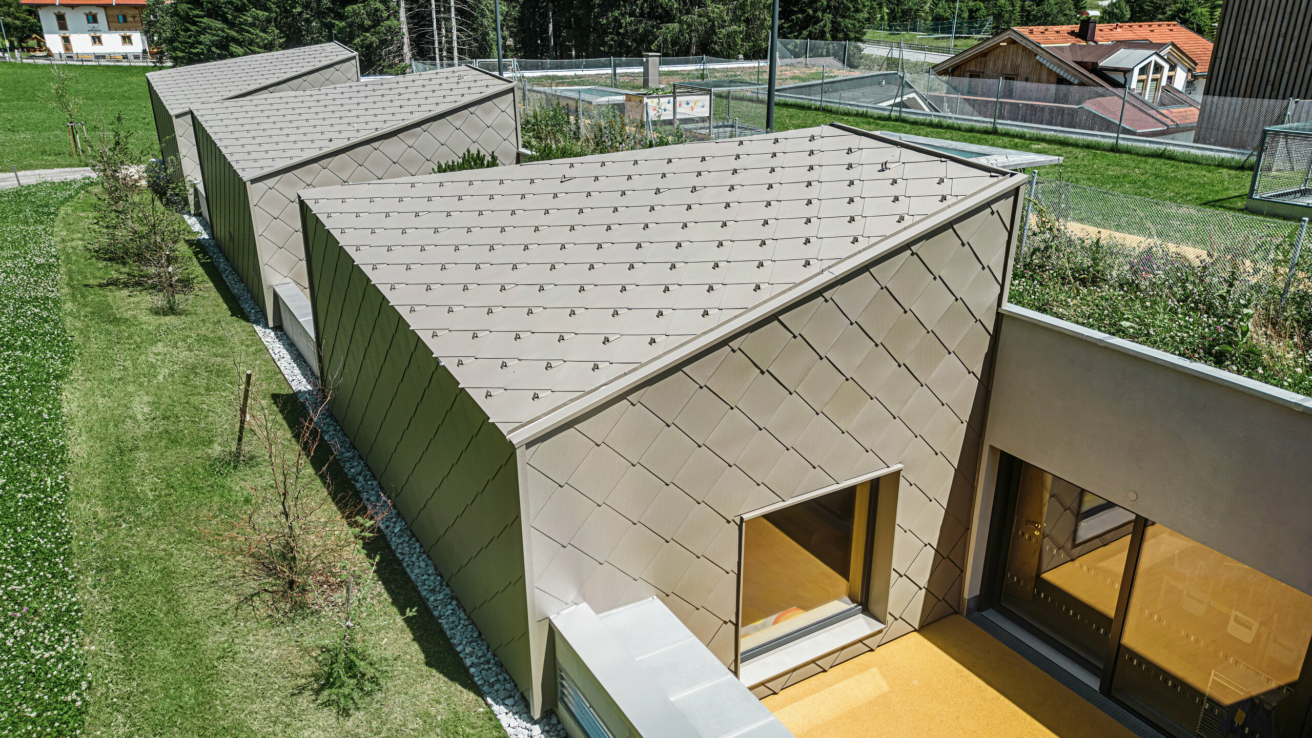 Bronze-farbene PREFA Dach und Wandrauten 44 × 44 bedecken das Dach und Teile der Fassade eines modernen Gebäudes in Gerlos, Zillertal. Das Gebäude verfügt über großzügige Fensterfronten und Terrassenbereiche. Im Hintergrund ist die ländliche Umgebung mit grünen Wiesen und einem bewaldeten Hügel zu sehen.