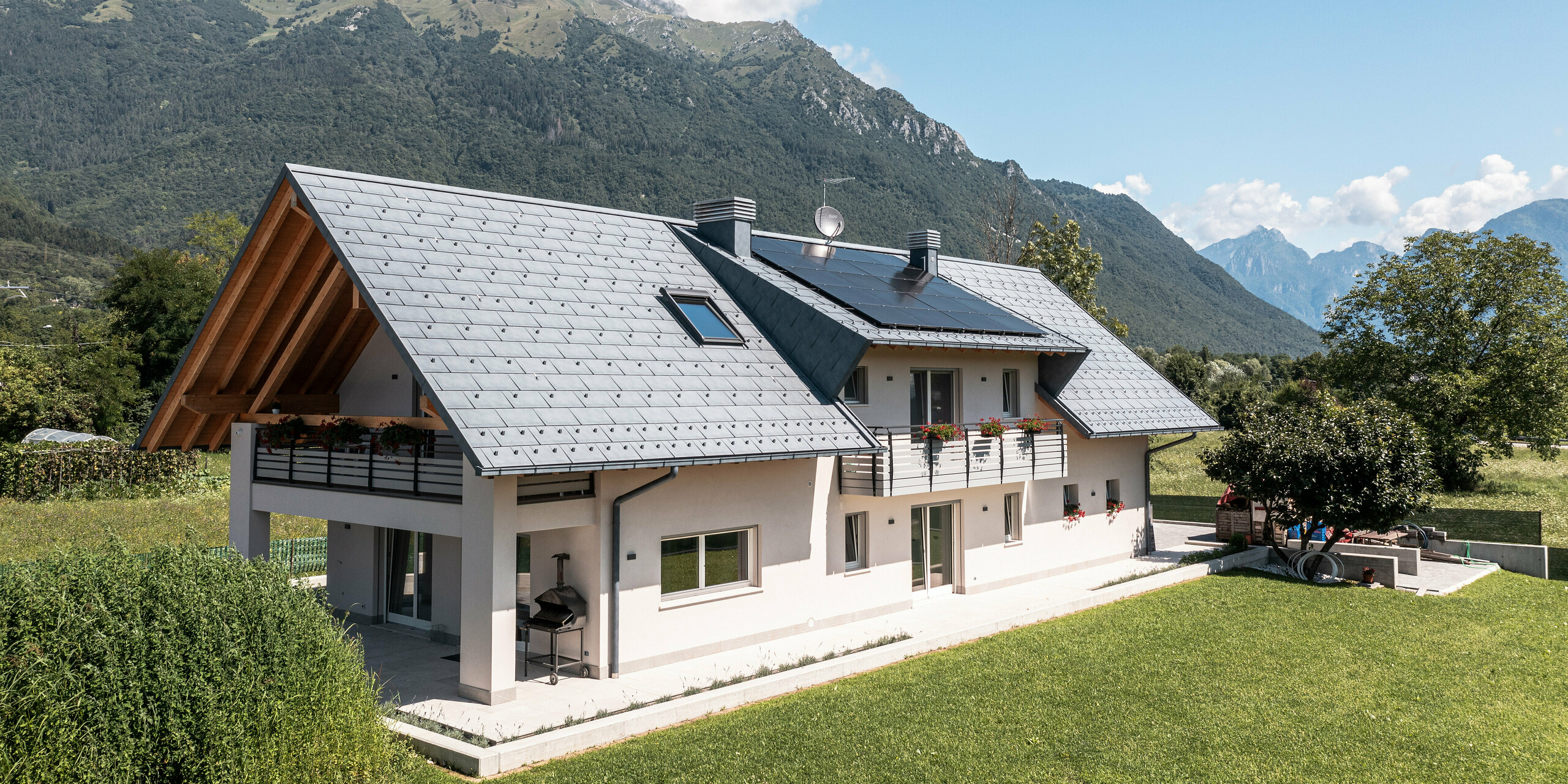 Drohnenaufnahme eines Hauses in Belluno, Italien, eingedeckt mit PREFA R.16 Dachplatten in P.10 anthrazit. Das Anwesen liegt in einer atemberaubenden Alpenlandschaft und kombiniert traditionelle Elemente wie freiliegende Holzgiebel mit modernen, langlebigen Baumaterialien, wodurch eine perfekte Symbiose aus Tradition und Moderne entsteht.