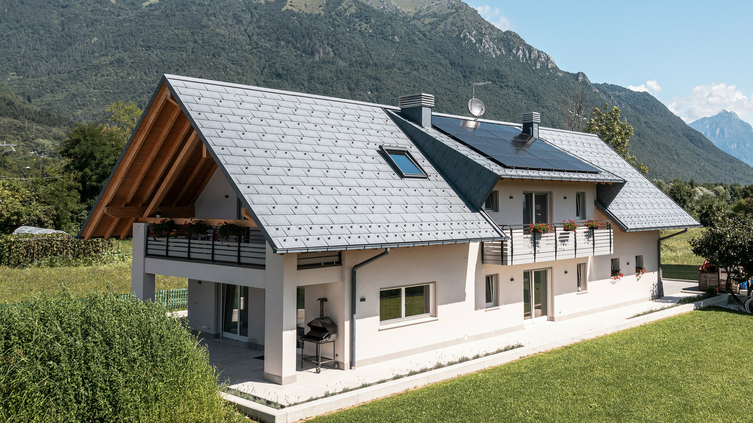 Drohnenaufnahme eines Hauses in Belluno, Italien, eingedeckt mit PREFA R.16 Dachplatten in P.10 anthrazit. Das Anwesen liegt in einer atemberaubenden Alpenlandschaft und kombiniert traditionelle Elemente wie freiliegende Holzgiebel mit modernen, langlebigen Baumaterialien, wodurch eine perfekte Symbiose aus Tradition und Moderne entsteht.