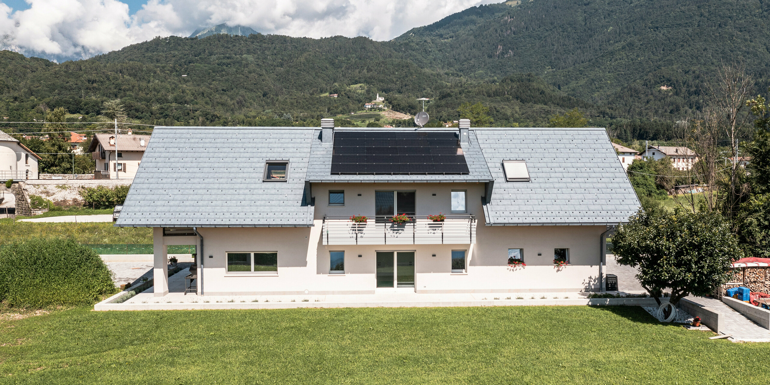 Idyllisches Einfamilienhaus in Bellun, ausgestattet mit PREFA R.16 Dachpaneelen in der Farbe Anthrazit, inmitten einer saftig grünen Wiese gelegen. Die klaren Linien der Architektur und das moderne Dach bilden einen wunderbaren Kontrast zu den traditionellen Landhäusern im Hintergrund und der umliegenden Berglandschaft.