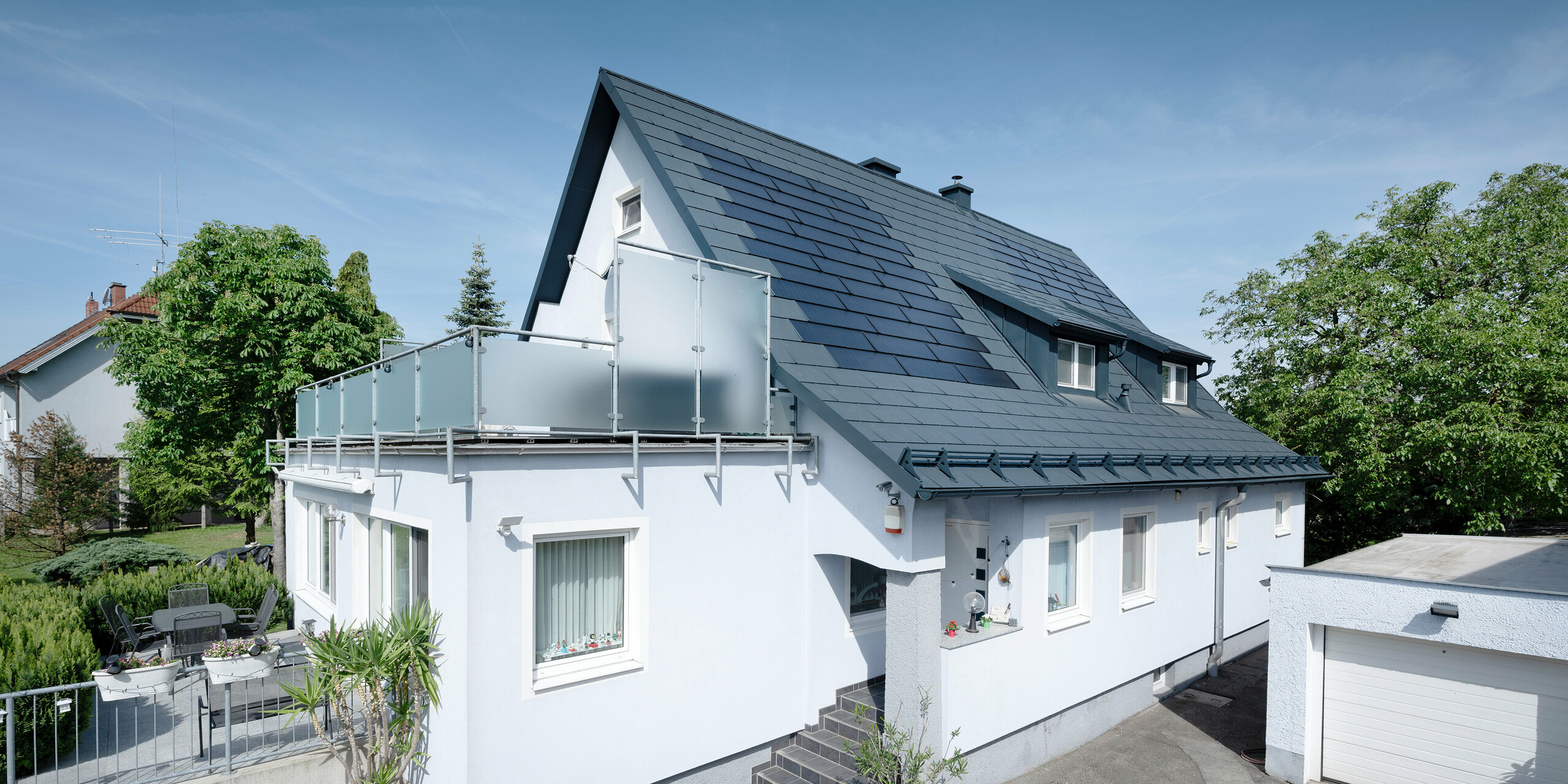 Dakrenovatie van een vrijstaand huis in Leonding  met PREFA-zonnedakpanelen in antraciet. Op de foto is het gerenoveerde huis van opzij te zien met een duidelijk zicht op het dak en de geïntegreerde zonnepanelen. Deze bieden een duurzame en energie-efficiënte manier om stroom op te wekken. In deze woning wekken de zonnepanelen een stroomopbrengst van 10,6 kWp op. De moderne, duurzame en weerbestendige PREFA-materialen dragen bij aan een esthetische en functionele dakconstructie die voldoet aan de nieuwste normen voor duurzaam bouwen.