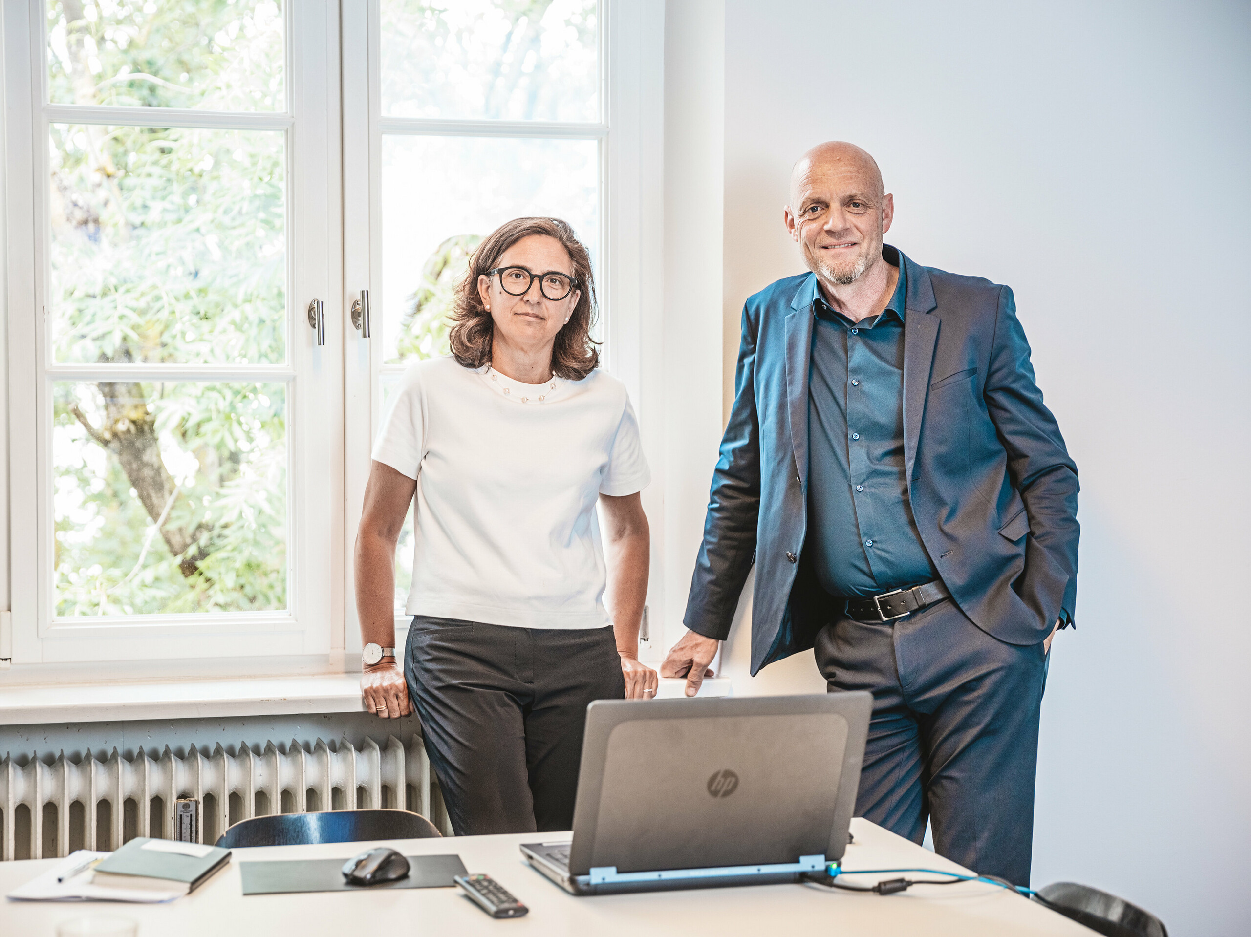 Patricia Ezcura und Gerold Heugenhauser von WWA Architekten aus München, die kreativen Köpfe hinter dem Wohnprojekt in Utting am Ammersee. Die beiden stehen in einem Büro hinter einem Tisch mit einem Laptop und Notizbüchern und vor einem großen Fenster. Ihre innovative Planung und Gestaltung des modernen Gebäudekomplexes am Schmucker-Areal mit nachhaltigen PREFA Produkten wie der Dachplatte R.16 zeichnet sich durch eine harmonische Integration in die Umgebung und eine ästhetisch ansprechende Architektur aus.