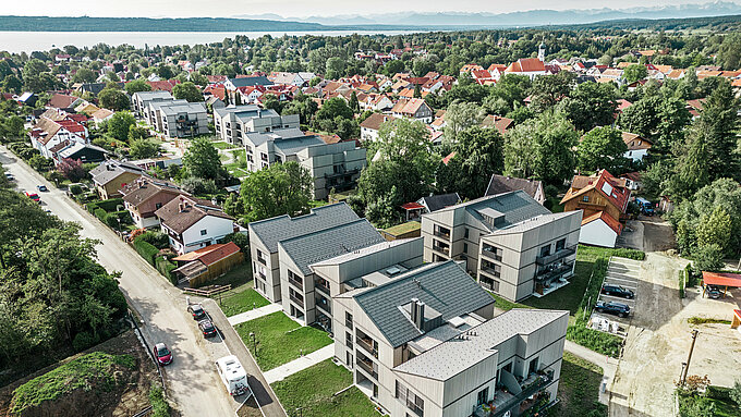 Panoramaaufnahme des großen Schmucker-Areals in Utting am Ammersee, das sich durch eine weitläufige und moderne Bebauung mit mehreren Mehrfamilienhäusern auszeichnet. Die sechs Baukörpern sind mit nachhaltigen und langlebigen PREFA Dachplatten R.16 in Dunkelgrau ausgestattet und fügen sich harmonisch in die Umgebung ein. Die Gebäude sind umringt von grünen Bäumen und im Hintergrund blitzt das Blau des Ammersees hervor. Die Architektur des gesamten Wohnprojektes besticht durch klare Linien und eine ästhetische Kombination aus Holzfassaden und hochwertigen Dachsystemen aus Aluminium, die sowohl Funktionalität als auch visuelle Eleganz bieten.