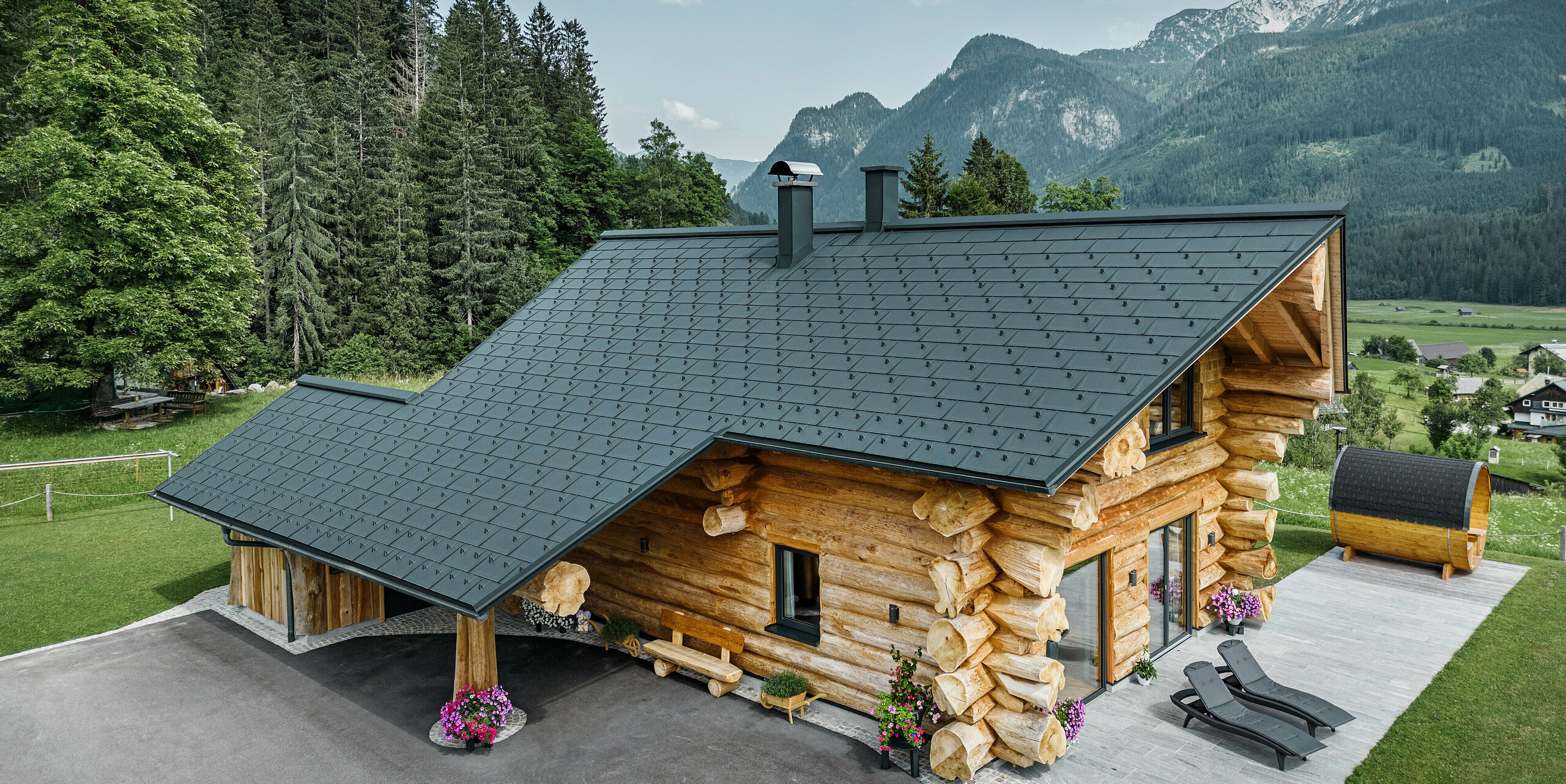 Schräge Ansicht eines malerischen Blockhauses in Gosau, Österreich, ausgestattet mit einem PREFA Dach aus anthrazitfarbenen Dachplatten R.16. Die Architektur kombiniert rustikales Blockhausflair mit modernen Elementen, umgeben von dichten grünen Wäldern und einer beeindruckenden Bergkulisse. Das Haus strahlt mit seiner ländlichen Eleganz und den präzise verlegten Dachelementen aus Aluminium, die für ihre Langlebigkeit und Witterungsbeständigkeit bekannt sind, eine einladende Atmosphäre aus.