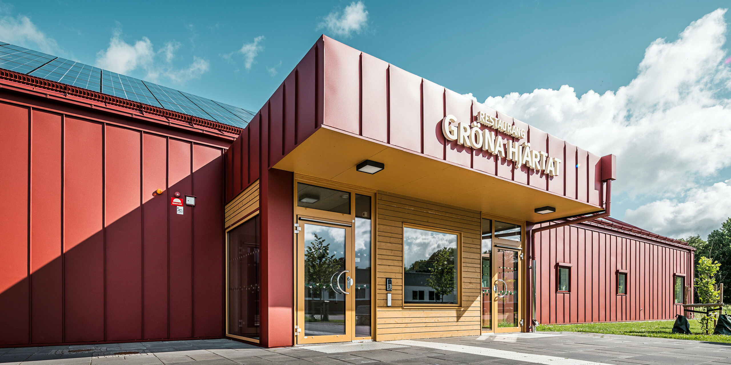 Nahaufnahme des Eingangsbereichs des Restaurants Gröna Hjärtat, der Schulkantine der Göingeskolan und Prästavångskolan in Broby, Schweden. Die Fassade und das Dach sind mit PREFALZ in Oxydrot verkleidet, was dem Gebäude eine markante, moderne Optik verleiht. Über dem Eingang prangt das Schild 'Restaurang Gröna Hjärtat'. Die Kombination aus einer langlebigen Aluminiumverkleidung und energieeffizientem Design, einschließlich einer auf dem Dach installierten Photovoltaikanlage, macht das Gebäude zu einem herausragenden Beispiel zeitgenössischer Architektur. Die PREFA Dachentwässerung ist passend in Oxydrot ausgeführt und trägt zum harmonischen Gesamtbild bei.