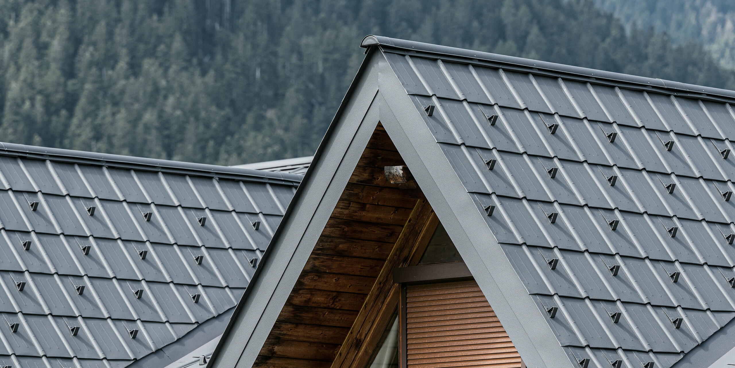 Nahaufnahme der Gaube auf dem Alpenhaus in Auronzo di Cadore, ausgestattet mit PREFA Ziegeln in Hellgrau. Die fein strukturierten Aluminiumpaneele bilden einen schönen Kontrast zum natürlichen Holz der Dachkonstruktion. Sie bieten einen hochwertigen Schutz vor dem alpinen Wetter und fügen sich nahtlos in die Bergwaldlandschaft ein.