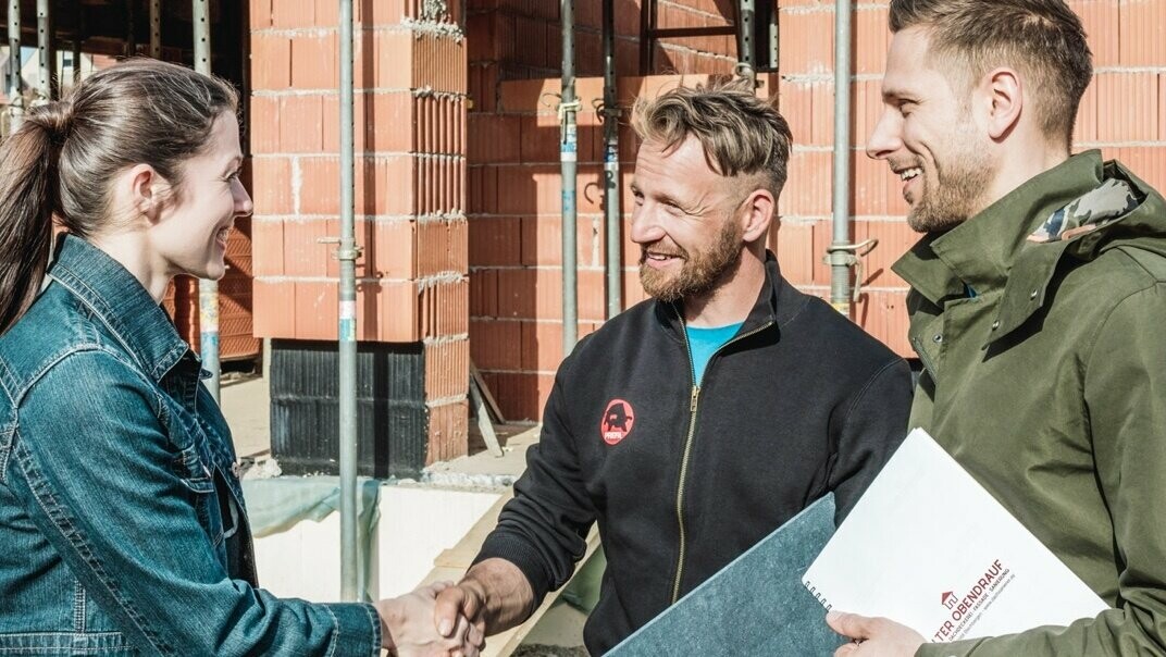 Een jong stel ontmoet de buurman op de bouwplaats van hun huis. Ze hebben het aanbod besproken en geaccepteerd. De blikslager houdt een PREFA-dakpaneel in zijn hand.