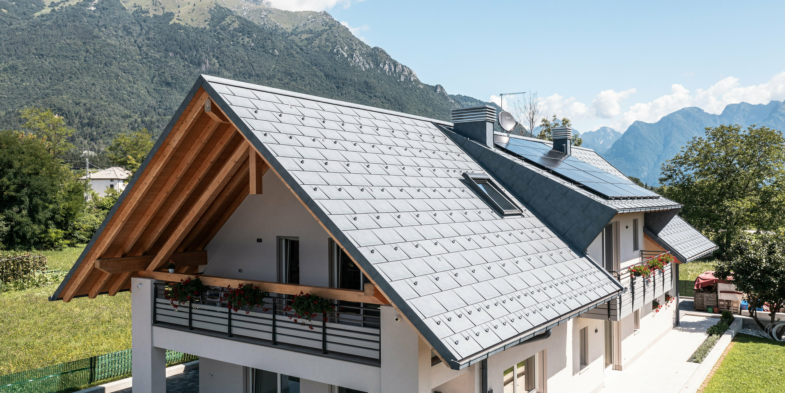 Idyllisches Einfamilienhaus in Bellun, ausgestattet mit PREFA R.16 Dachpaneelen in der Farbe Anthrazit, inmitten einer saftig grünen Wiese gelegen. Die klaren Linien der Architektur und das moderne Dach bilden einen wunderbaren Kontrast zu den traditionellen Landhäusern im Hintergrund und der umliegenden Berglandschaft.