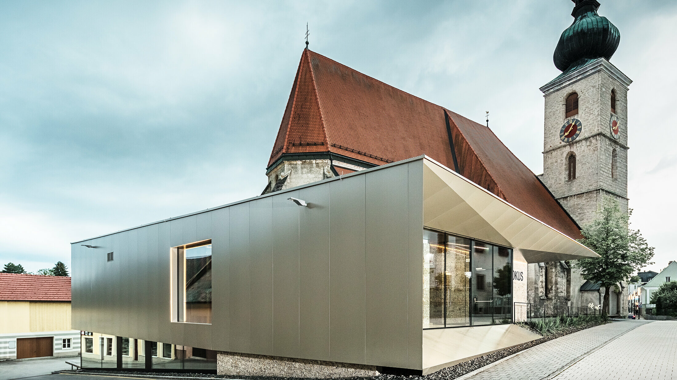 De opvallende bronskleurige gevel met composietplaten van het parochiecentrum in Sierning strekt zich uit rond de kleine kerk op de achtergrond