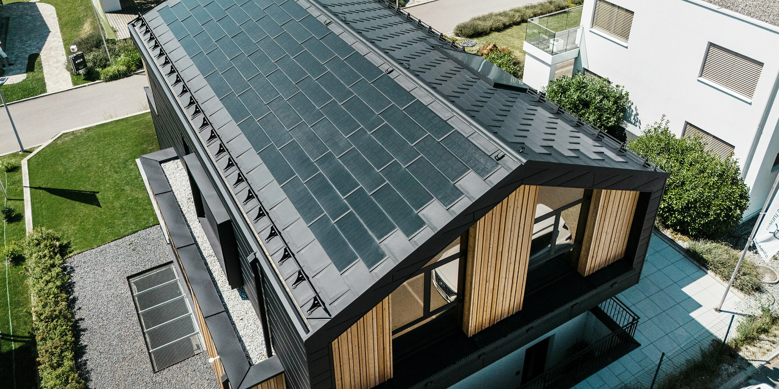 Vogelperspektive auf das Musterhaus Autark im Fertighauszentrum Blaue Lagune, Vösendorf (AT), mit robustem PREFA Aluminiumdach. Das Dach ist mit PREFA Solardachplatten und Dachplatten R.16 in P.10 Schwarz eingedeckt. Die integrierte PV-Anlage sorgt für nachhaltige Energiegewinnung durch die Kraft der Sonne. Die Holzverschalung an der Giebelseite rundet das zeitgemäße Erscheinungsbild des Hauses ab.