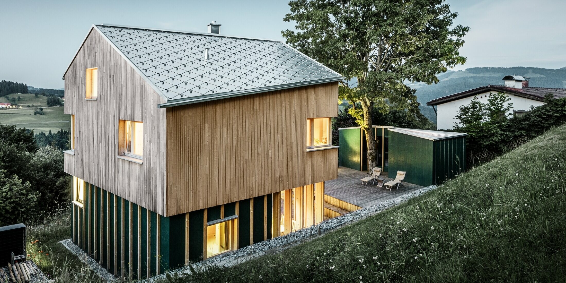 Ein Einfamilienhaus in Hanglage wurde mit der PREFA Dachraute 44x44 in Naturblank eingedeckt. Das langlebige Aluminiumdach harmoniert perfekt mit der hellen Holzfassade. Die Fenster des Gebäudes strahlen Licht und Wärme aus und kontrastieren mit der abendlichen Dämmerung im Hintergrund. 