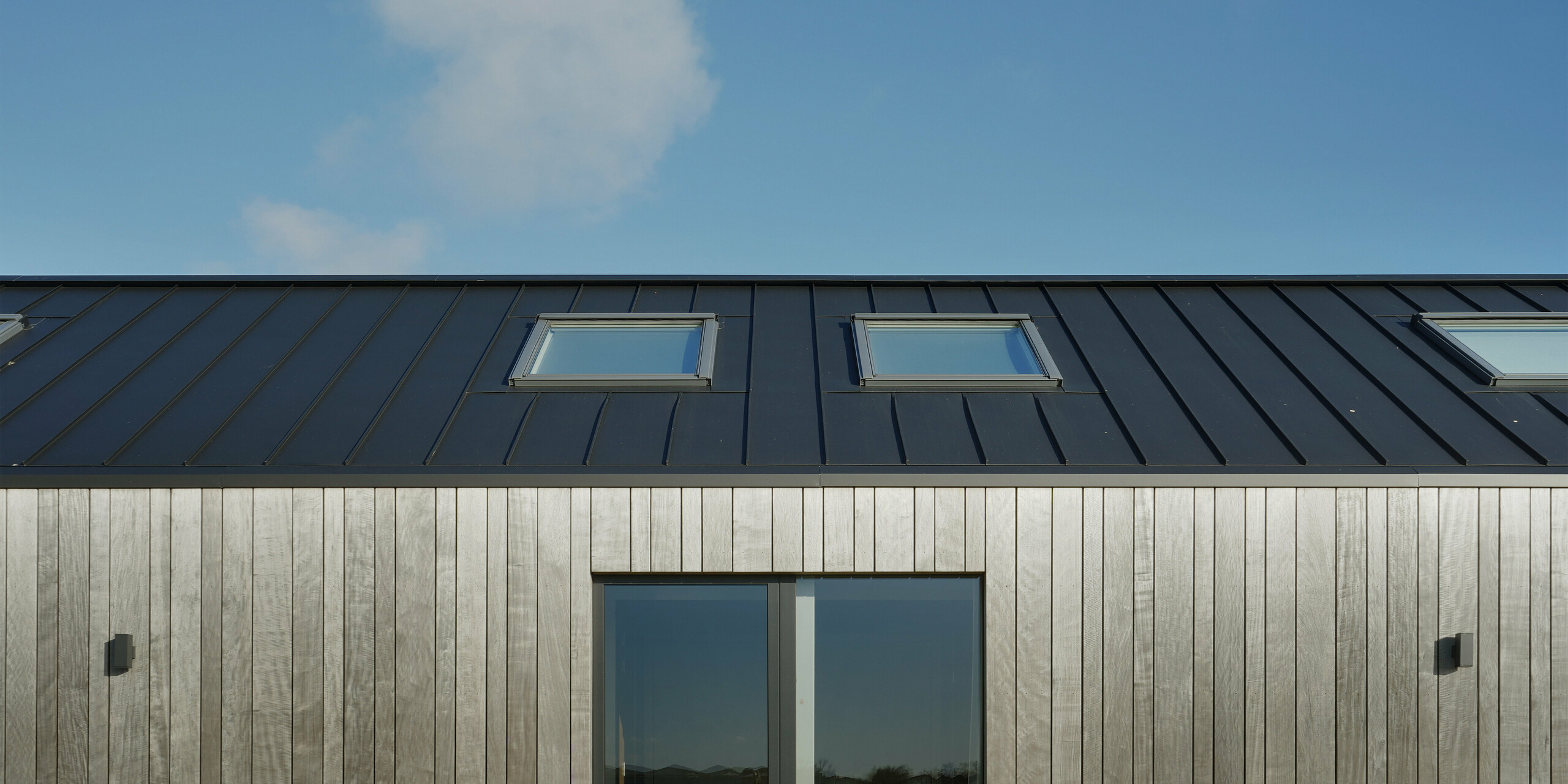 Nahaufnahme der PREFALZ Winkelstehfalz-Eindeckung in Schwarzgrau eines modernen Einfamilienhauses in Uithoorn, Niederlande. Das Bild hebt die präzise Integration der Dachfenster in das hochwertige Aluminiumdach hervor. Die Kombination aus robusten, wetterbeständigen Materialien und natürlichen Holzelementen an der Fassade unterstreicht das nachhaltige und zeitgemäße Design dieser Wohnscheune.