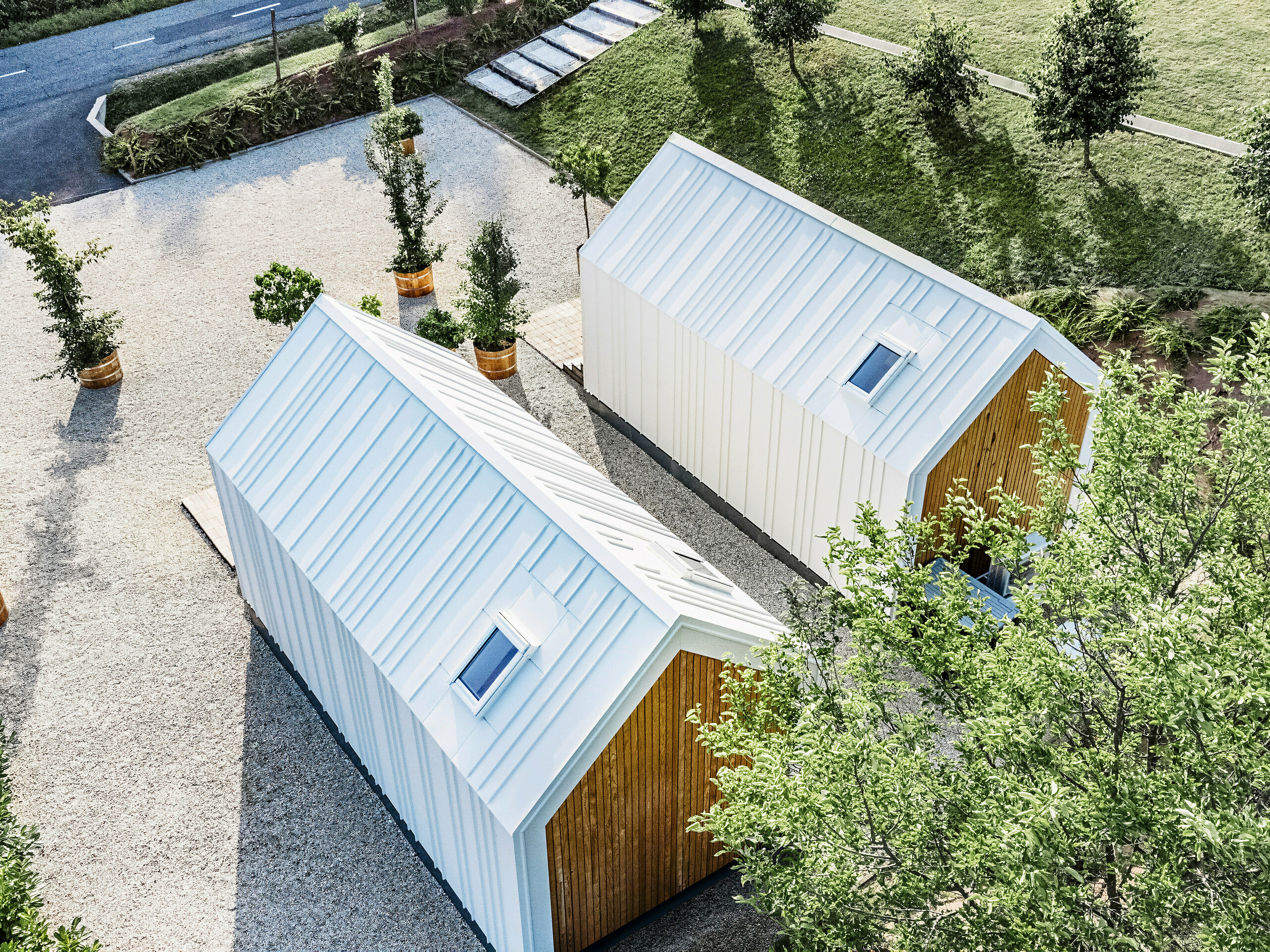 Vogelperspektive auf die Rückseite der beiden Tiny Houses 'Pri Momi' in Kančevci, Slowenien. Die exklusiven Mikrohäuser sind mit einer robusten PREFALZ Dach- und Fassadenbekleidung in der Farbe P.10 Reinweiß sowie Naturholz ausgestattet. Die weiße Gebäudehülle aus hochwertigem und langlebigem Blech wurde in einer Stehfalzdeckung ausgeführt. Die minimalistische Architektur fügt sich harmonisch in die umgebende Grünanlage und die gepflasterten Wege ein. Das Bild zeigt auch die leicht versetzte Anordnung der Gebäude, die in Holzrahmenbauweise errichtet wurden.