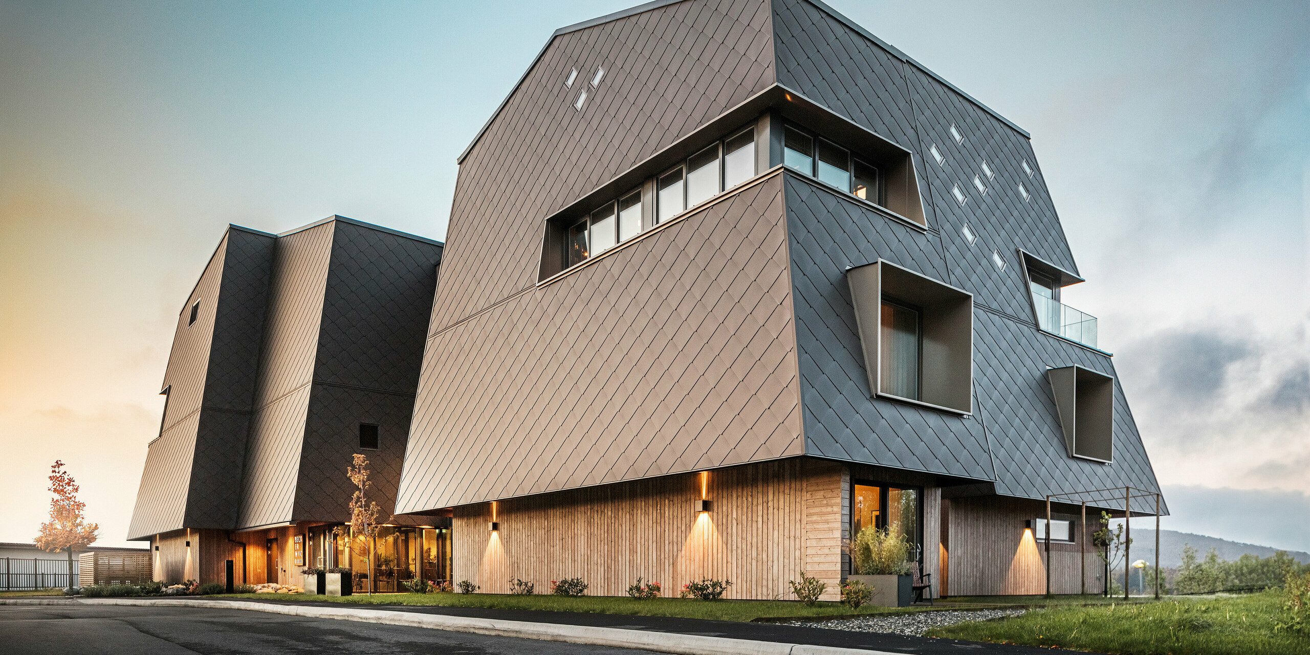 Blick auf den Eingangsbereich des Hotels Beckerwyc in Bekkjarvik, Norwegen, bei Dämmerung. Die Fassade ist mit PREFA Wandrauten 44 × 44 in Nussbraun verkleidet, was dem Gebäude ein modernes und zugleich beeindruckendes Aussehen verleiht. Die Beleuchtung betont die architektonischen Details und die außergewöhnliche Form des Gebäudes. Die Kombination aus futuristischem Design und natürlicher Farbgebung fügt sich harmonisch in die Umgebung ein, was durch die angenehmen Lichtverhältnisse zusätzlich betont wird.