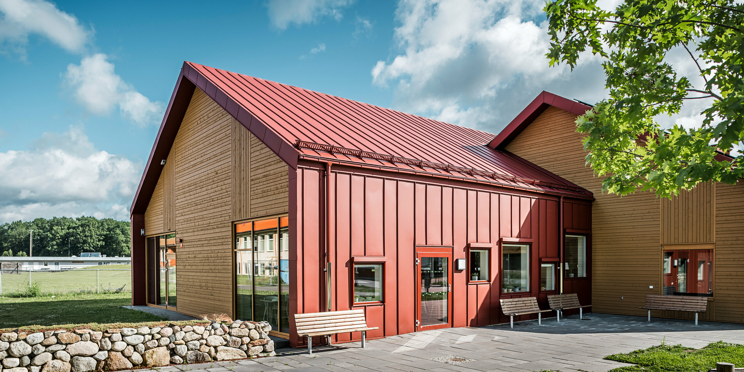 Außenansicht der Schulkantine Restaurang Gröna Hjärtat in Broby, Schweden. Das Bild zeigt Sitzbänke vor dem Gebäude, das eine markante Kombination aus oxydrotem PREFALZ und Holz an der Giebelseite aufweist. Die architektonische Gestaltung betont klare Linien und den harmonischen Kontrast zwischen den roten Aluminiumflächen und dem warmen Holzton. Im Vordergrund sind Steinmauern und Grünflächen zu sehen, die das moderne Design des Gebäudes umrahmen. Die Umgebung und das Gebäude verschmelzen zu einem einladenden und gut durchdachten Raum.