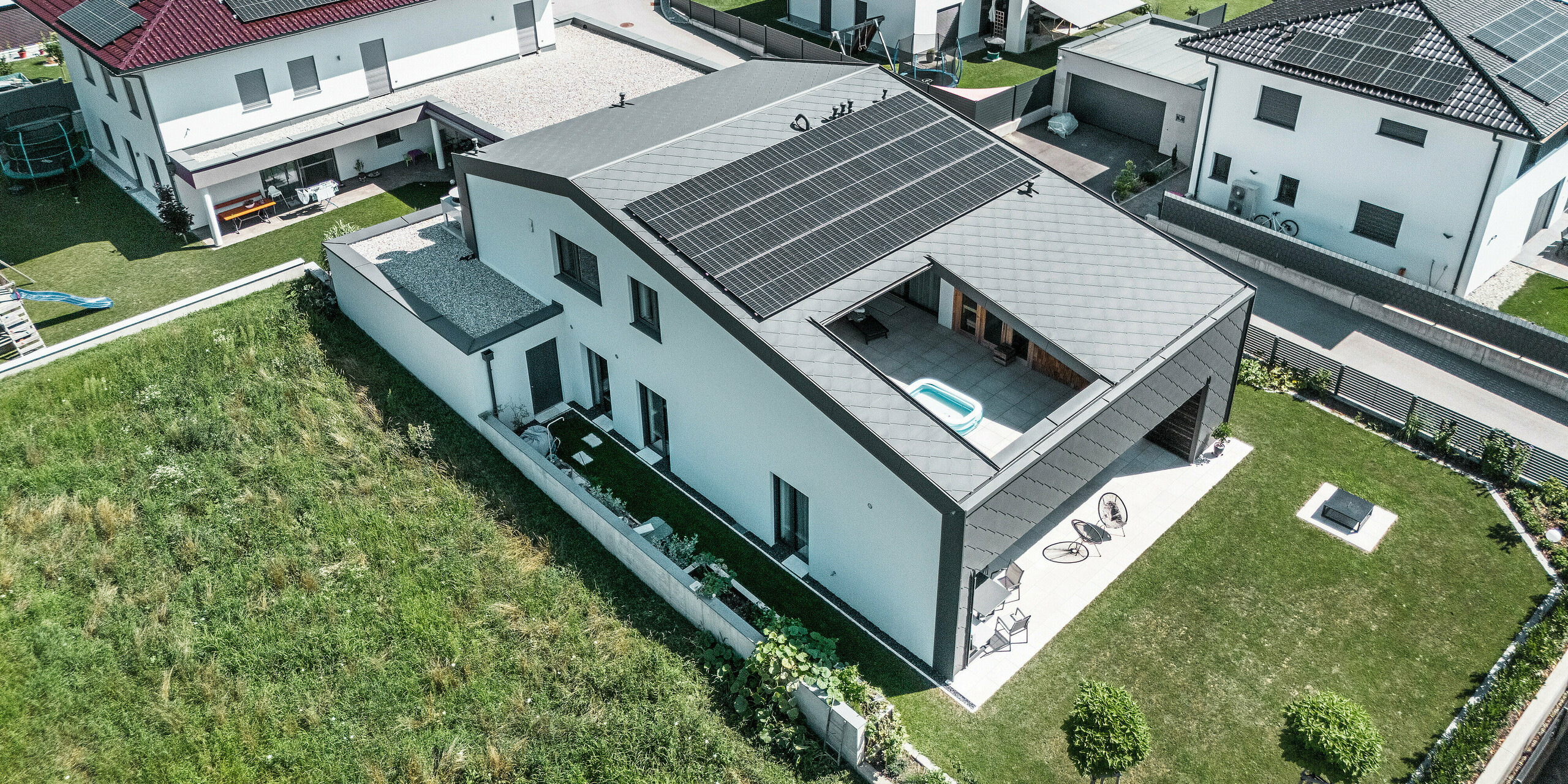 Drohnenaufnahme der Rückseite eines neuzeitlichen Einfamilienhauses in Aschach mit großzügigem Garten. Fassade und Dach sind mit dunkelgrauen PREFA Dach- und Wandrauten 44 × 44 eingedeckt. Die Dachfläche ist mit einer Photovoltaikanlage und einem Atrium ausgestattet, das zusätzlichen Wohnraum im Freien bietet. Die überdachte Terrasse im Erdgeschoss und das Atrium verleihen dem Haus eine offene, lichtdurchflutete Atmosphäre. Die Kombination aus Aluminiumverkleidung und weißer Putzfassade sorgt für ein harmonisches, modernes Gesamtbild.