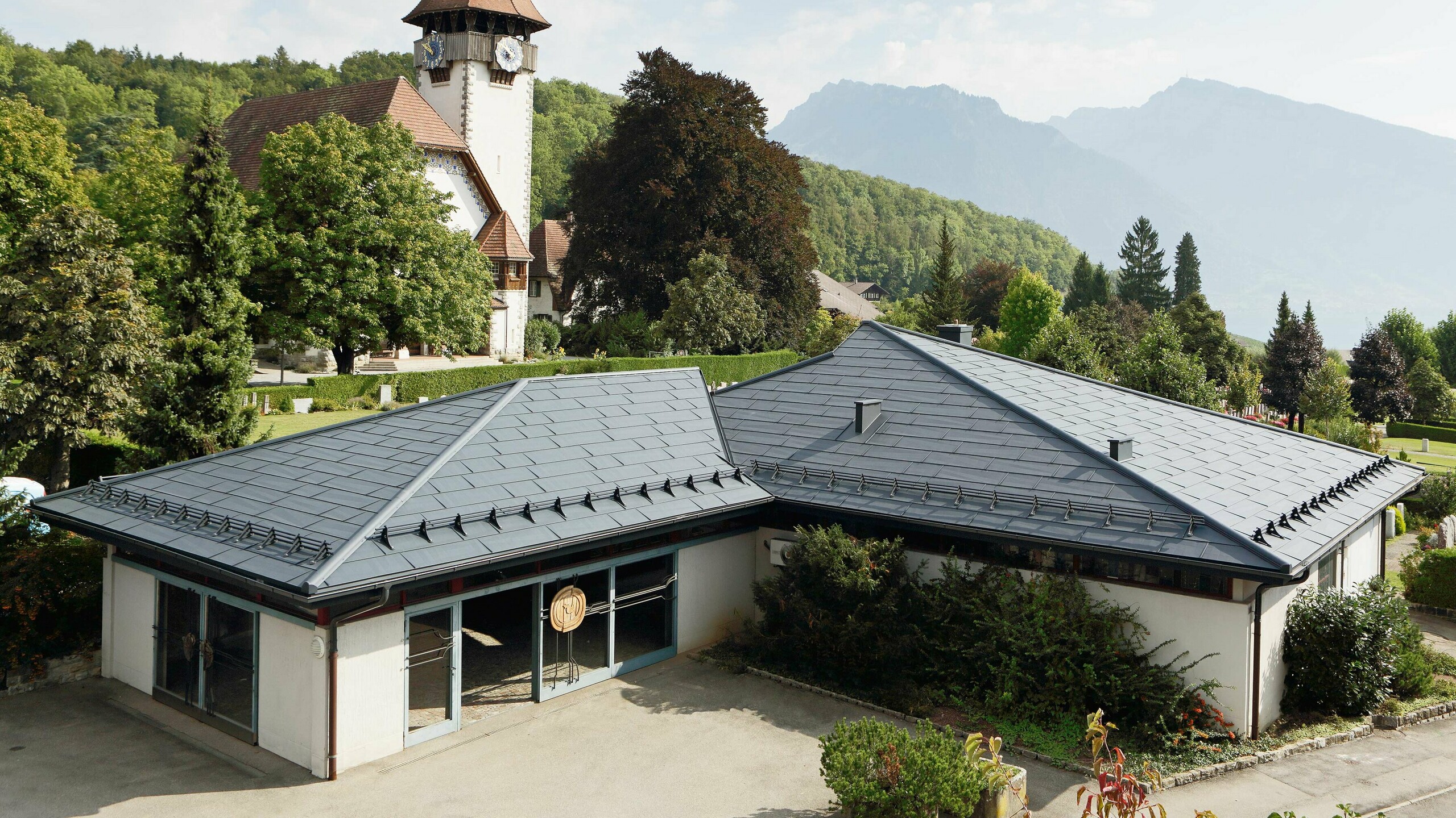 Rouwcentrum met FX.12 dakpanelen van PREFA in antraciet, op de achtergrond staat een kerk en daarnaast bevindt zich een kerkhof.