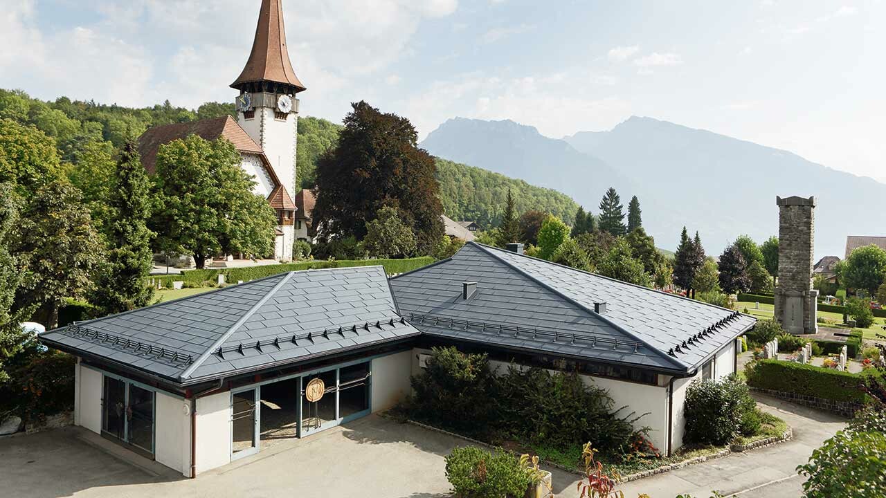 Rouwcentrum met FX.12 dakpanelen van PREFA in antraciet, op de achtergrond staat een kerk en daarnaast bevindt zich een kerkhof.