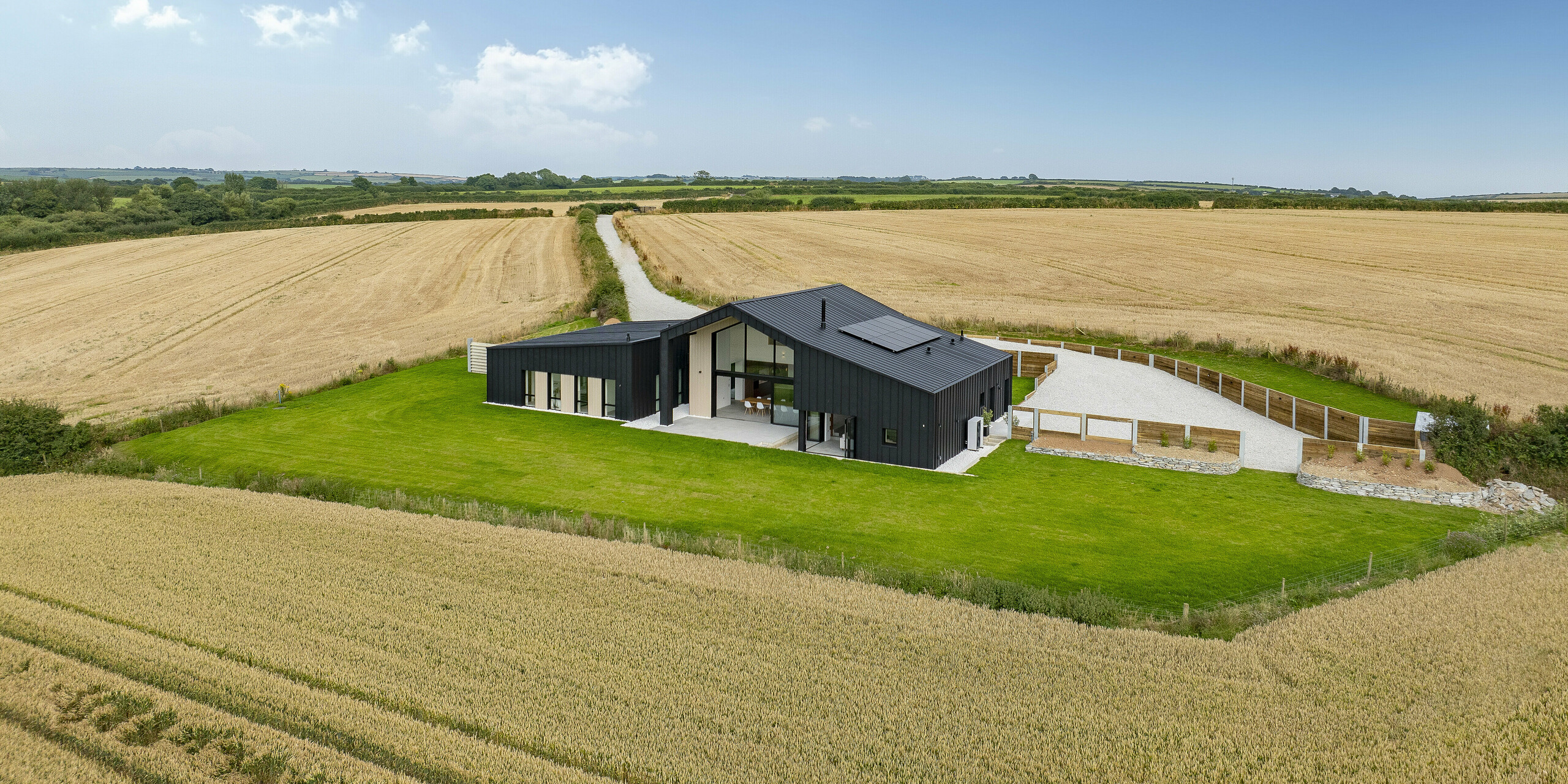 Panoramablick von ‚The Hide‘ in Cornwall, eingebettet in weite Felder. Vor dem Haus ist eine gepflegte Grünfläche zu sehen. Das moderne Einfamilienhaus beeindruckt mit einer anthrazitfarbenen PREFALZ Aluminiumhülle in der bewährten P.10 Beschichtung. Das Dach und die Fassade harmonieren perfekt mit der natürlichen Umgebung. Die klare Stehfalzoptik verleiht dem Gebäude Eleganz und Beständigkeit.