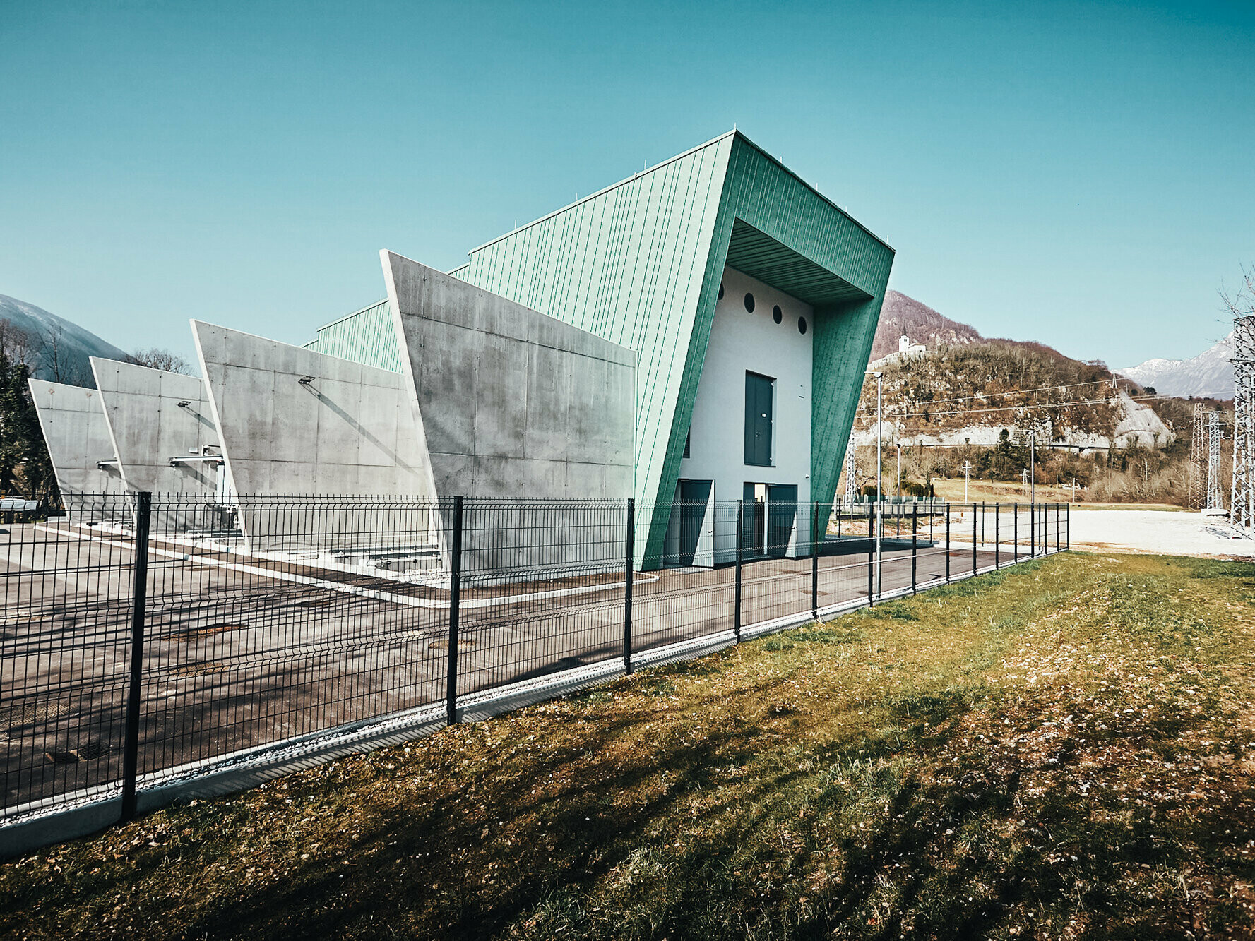 Seitenansicht des Umspannwerks samt Stirnseite und Betontrennwänden, eingezäunt und mit Bergen im Hintergrund.