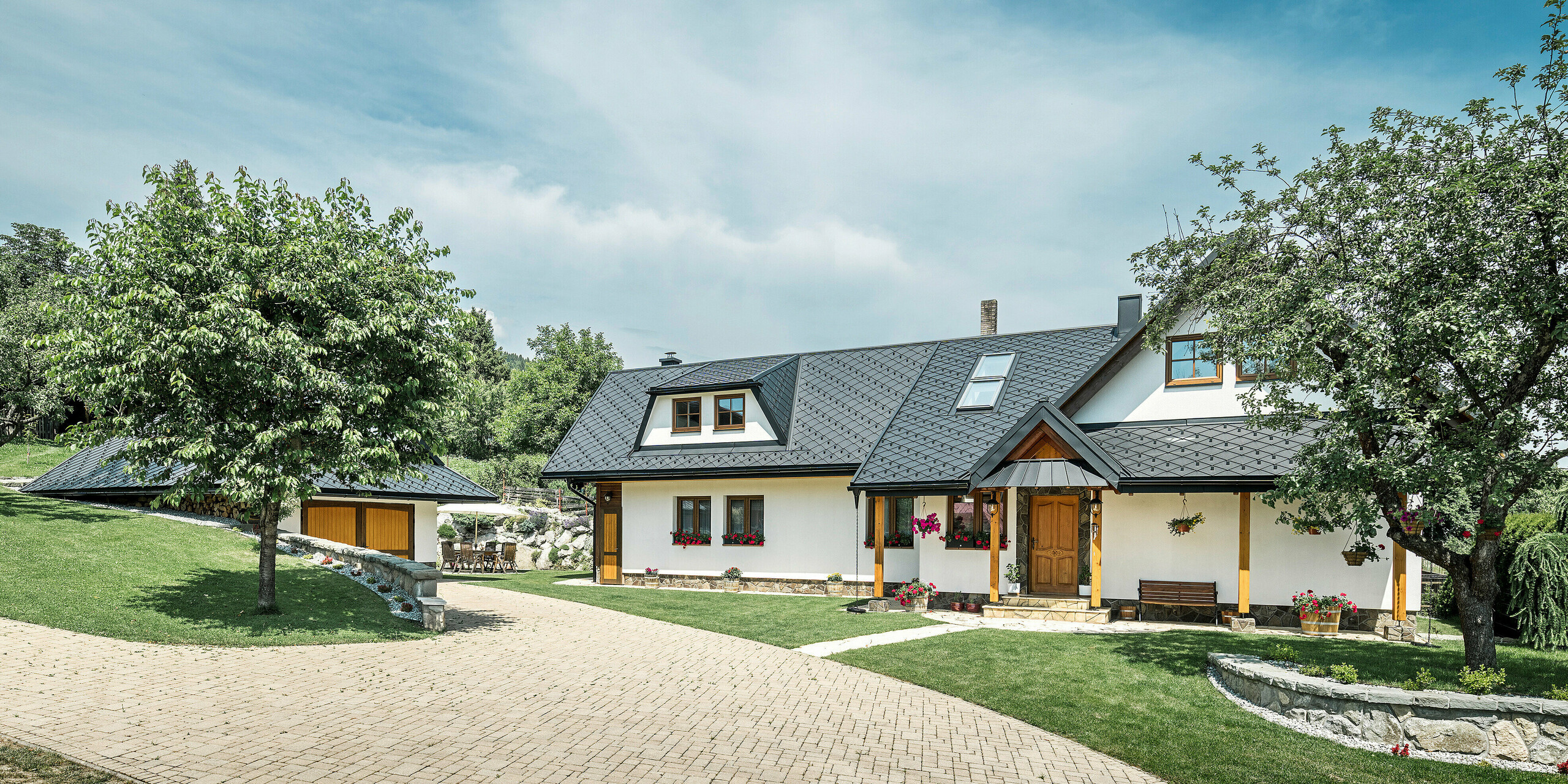 Zufahrt und Eingang eines traditionellen Einfamilienhauses in Stara Bystrica, Slowakei. Das Gebäude ist mit PREFA Dachrauten 29 × 29 in P.10 Schwarz gedeckt. Die Aluminiumrauten sorgen für robusten und langlebigen Schutz. Die gepflegte Pflastereinfahrt und die umgebende grüne Landschaft, mit einem Baum im Vordergrund, ergänzen das harmonische Gesamtbild des Hauses.