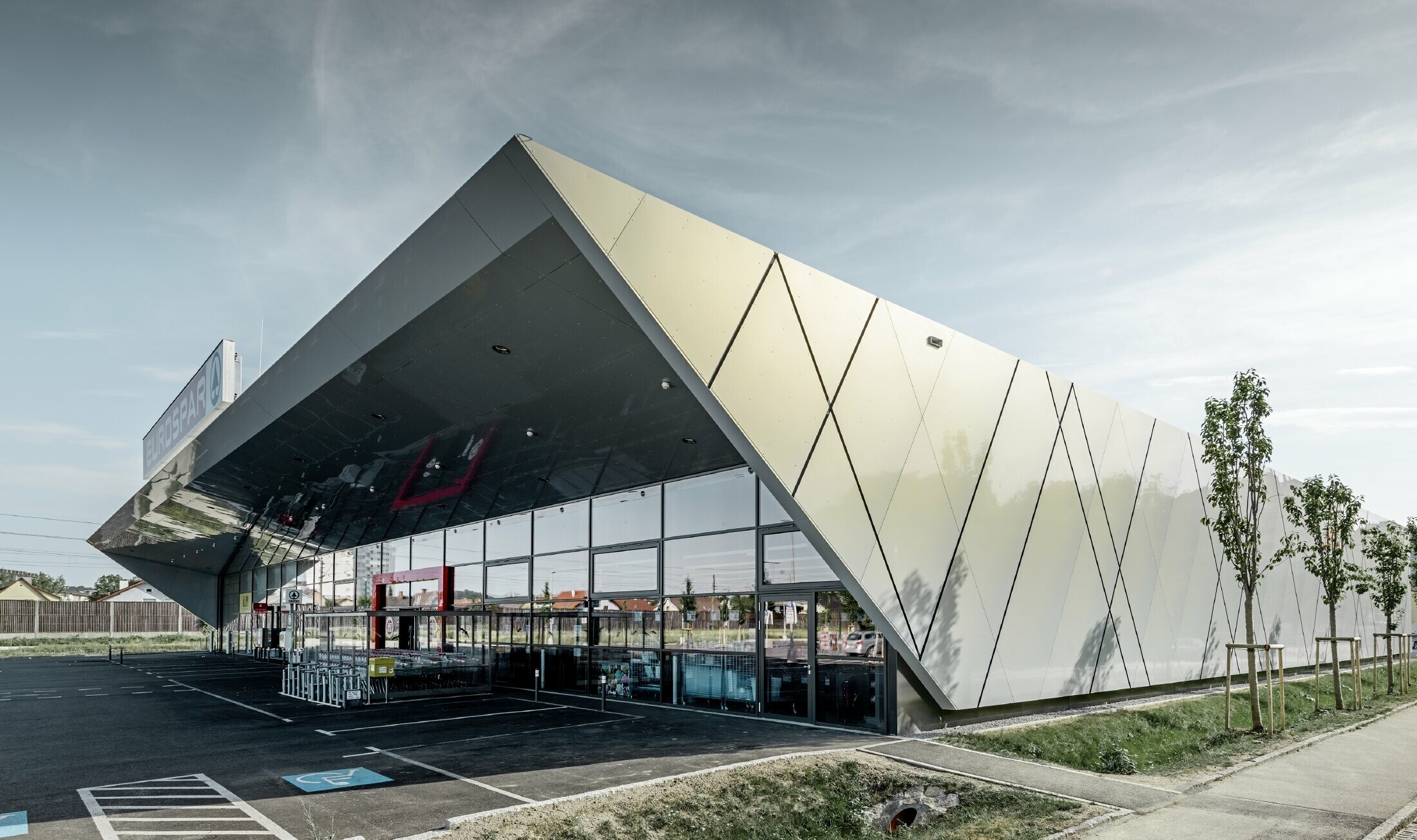 Entreegedeelte van het nieuwe Eurospar filiaal in Wels met het moderne gevelpaneel van PREFA in zilver goud; afhankelijk van de kijkhoek glinsteren de aluminium composietplaten in de zon in verschillende kleuren.