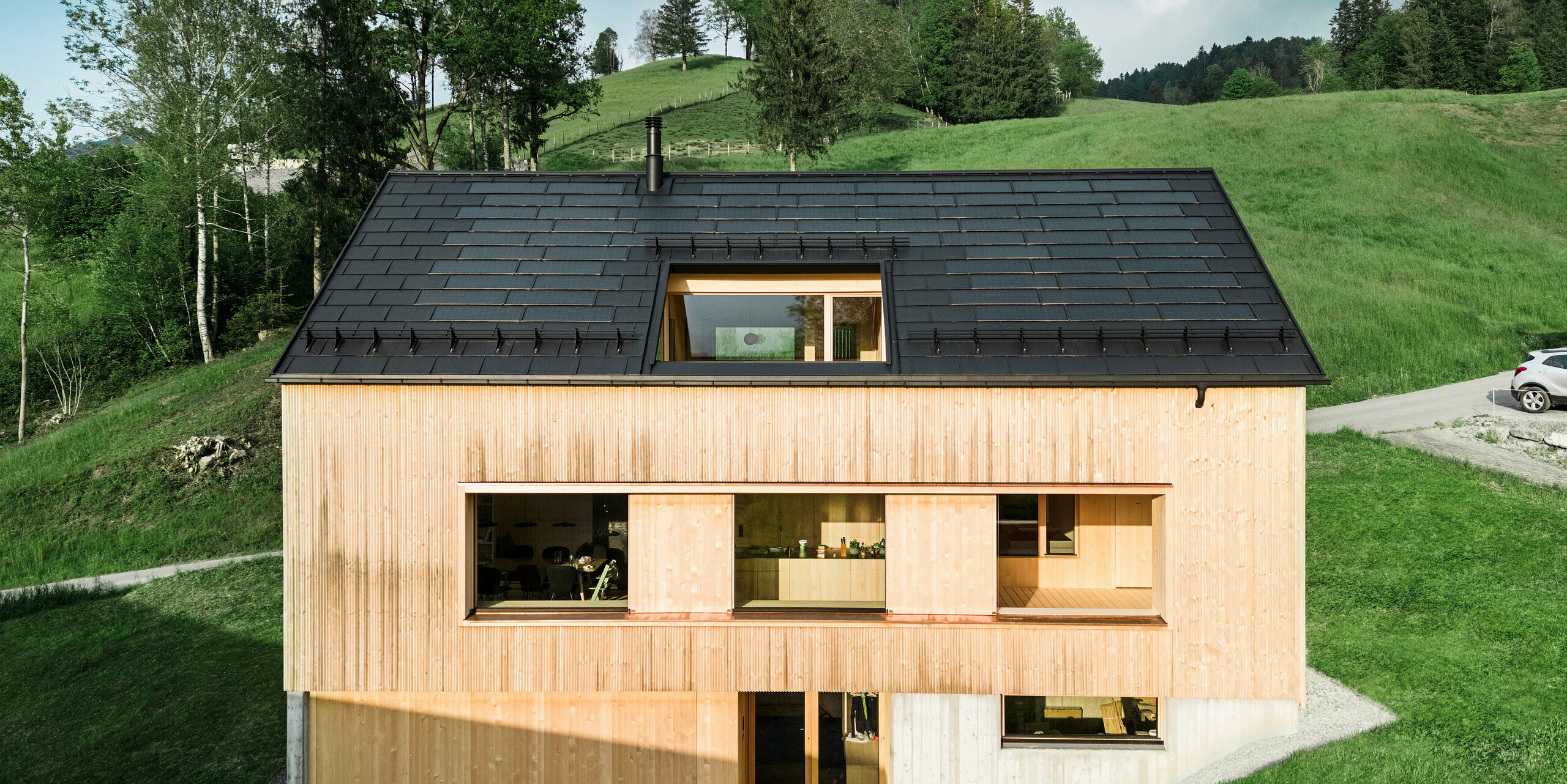 Ökologisch und ästhetisch: Das Bild zeigt ein modernes Ökohaus in Egg, ausgestattet mit der innovativen PREFA Solardachplatte. Das tiefschwarze, robuste Aluminiumdach, ausgeführt mit PREFA Dachplatten R.16, verschmilzt optisch mit der natürlichen Umgebung und unterstreicht die klaren Linien der zeitgenössischen Holzarchitektur. Die integrierten Solarmodule fügen sich diskret ins Dachbild ein und demonstrieren eine nachhaltige Bauweise, die Energieeffizienz und Design miteinander verbindet. Die innovative PREFA Solardachplatten schaffen eine Symbiose aus Funktionalität und Design. Dieses nachhaltige Wohnkonzept vereint hochwertige Aluminiumdachplatten mit fortschrittlicher Solartechnik und fügt sich nahtlos in die idyllische Berglandschaft ein, während es gleichzeitig den ökologischen Fußabdruck des Hauses minimiert.