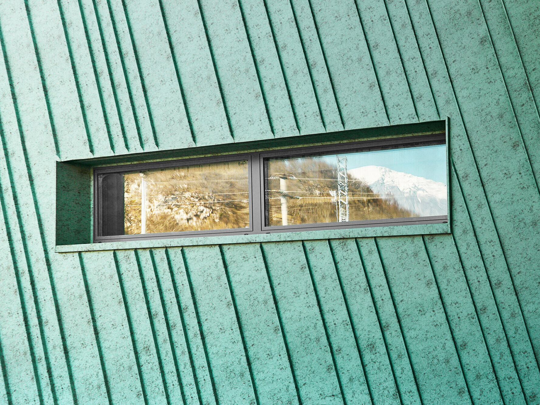 Detail mit leicht schräg verlaufender Fassade mit kleinem, länglichem Fenster, in dem sich Berge und elektrische Leitungen widerspiegeln. Seitenansicht auf Fassade und Glasfront mit Eingang.