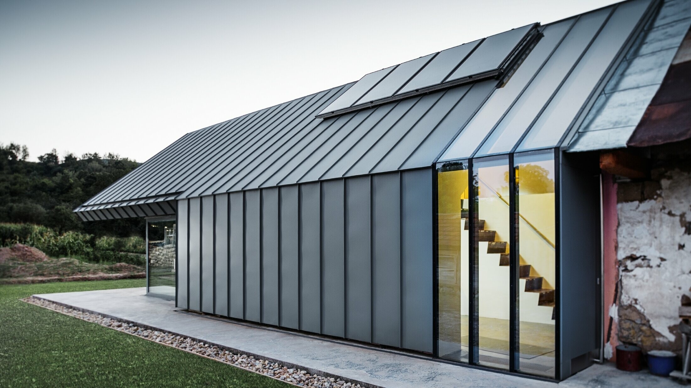 Modern architectenbureau/woning gebouwd tegen een oude boerderij, bekleed met staande naden en gebogen staande naden van PREFA in lichtgrijs